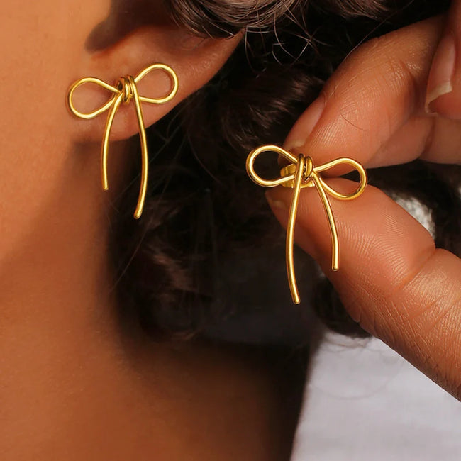Aferando anti-tarnish gold ribbon bow earrings worn on the ear, with a second earring held between fingers